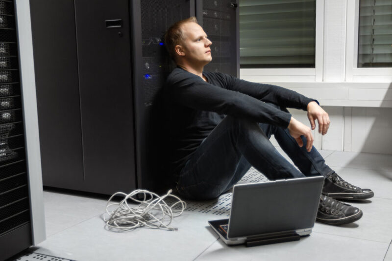 Frustrierter auf dem Boden vor einem Server sitzender IT Techniker Hochverfügbarkeit in der IT: Frustrierter auf dem Boden vor einem Server sitzender IT Techniker